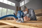 Một người mặc đồ bảo hộ đang sử dụng máy phay trên tấm ván gỗ trong xưởng.