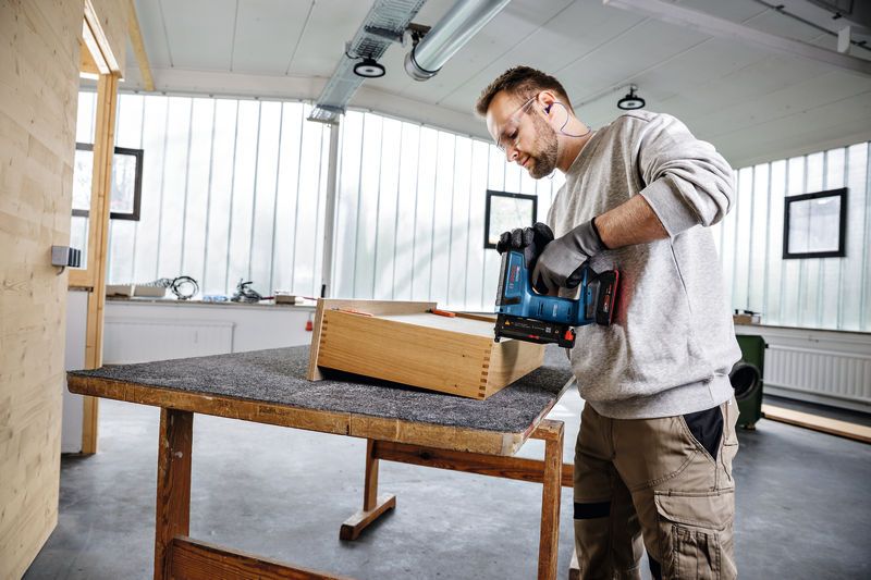 Người đeo thiết bị an toàn đang sử dụng súng bắn đinh không dây vào hộp gỗ trong xưởng.