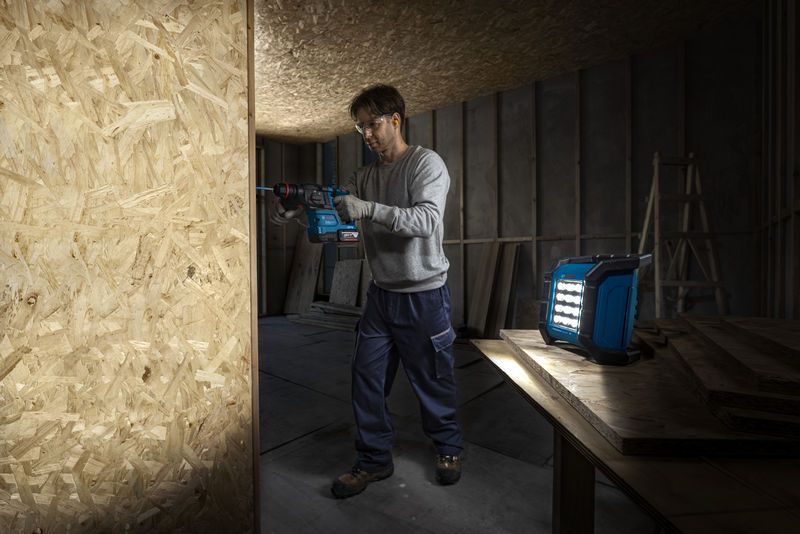 Một người mặc đồ bảo hộ đang khoan vào tấm gỗ gần đèn công trường không dây.