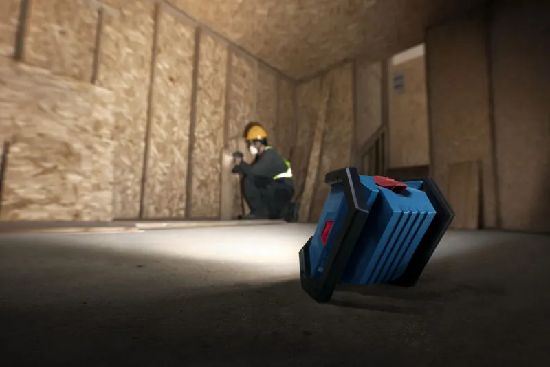 Một người mặc đồ bảo hộ đang lắp đặt vách thạch cao, được chiếu sáng bằng đèn công trường không dây.