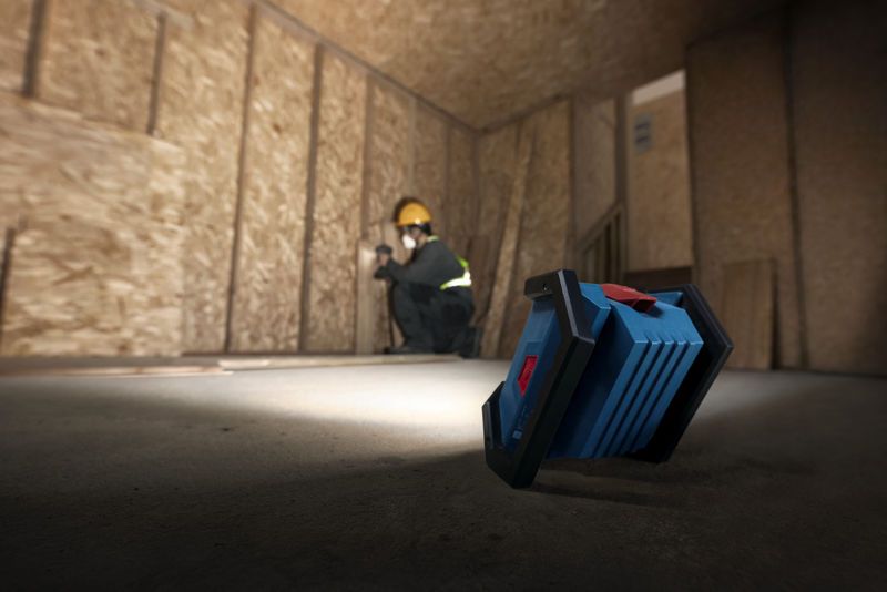 Một người mặc đồ bảo hộ đang lắp đặt vách thạch cao, được chiếu sáng bằng đèn công trường không dây.