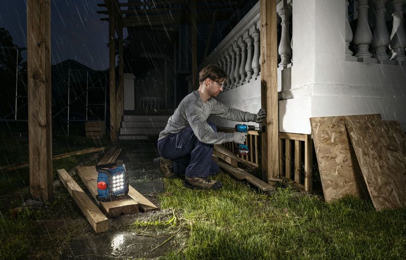 Một người mặc đồ bảo hộ đang khoan trụ gỗ vào ban đêm với đèn công trường không dây ở gần đó.