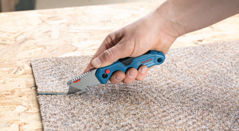 Một người đang dùng dao rọc giấy cắt thảm trên một bề mặt gỗ.