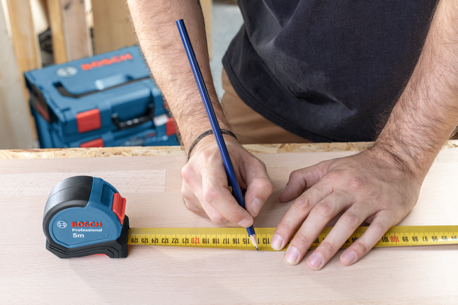 Một người đang dùng thước dây và bút chì để đánh dấu số đo trên tấm gỗ.