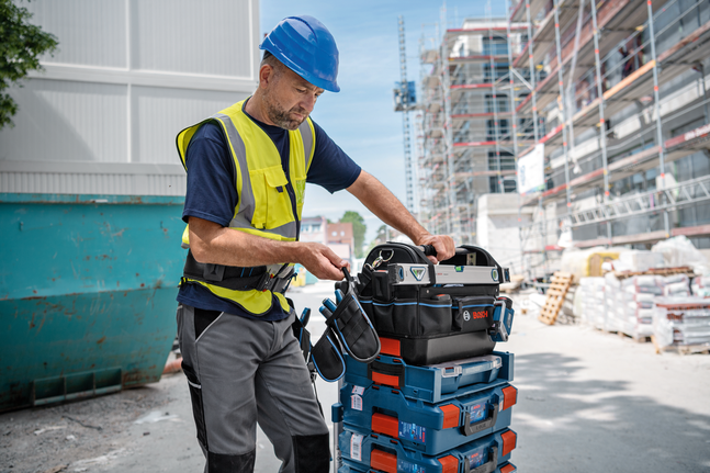 Một người mặc trang bị bảo hộ đang sắp xếp túi dụng cụ lên các hộp dụng cụ xếp chồng lên nhau tại công trường xây dựng.