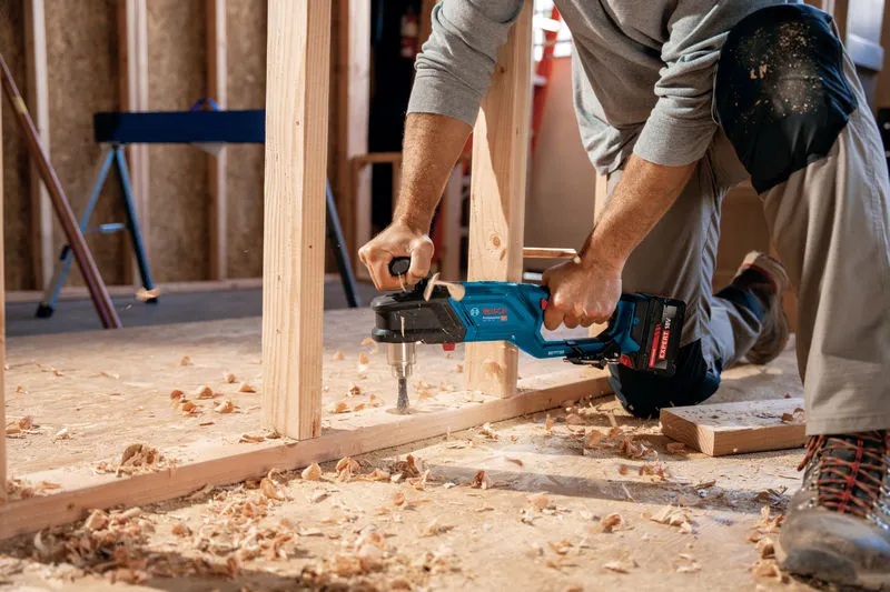 Một người mặc đồ bảo hộ đang khoan khung gỗ bằng máy khoan góc không dây.