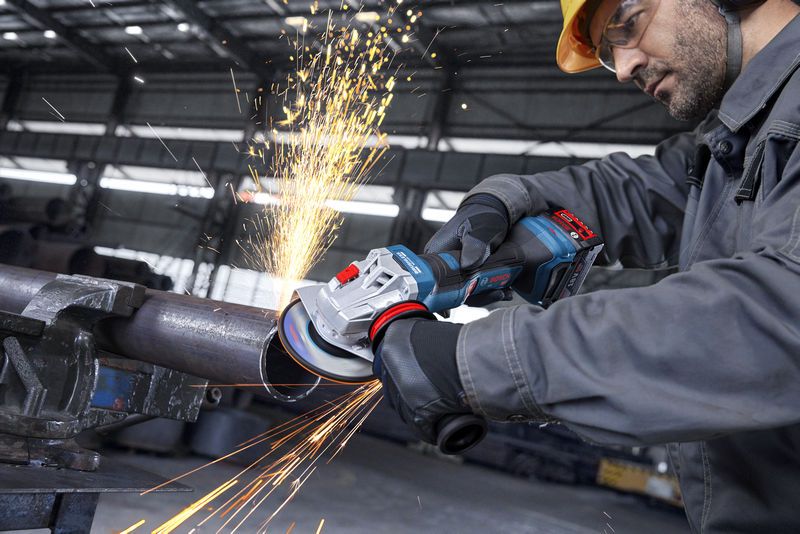 Một người đàn ông mặc đồ bảo hộ đang dùng máy mài góc để mài một ống kim loại, tạo ra tia lửa.