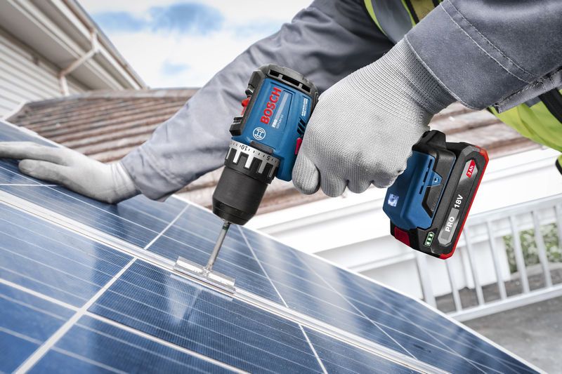 Một người mặc đồ bảo hộ đang cố định giá đỡ vào tấm pin mặt trời bằng máy khoan không dây.