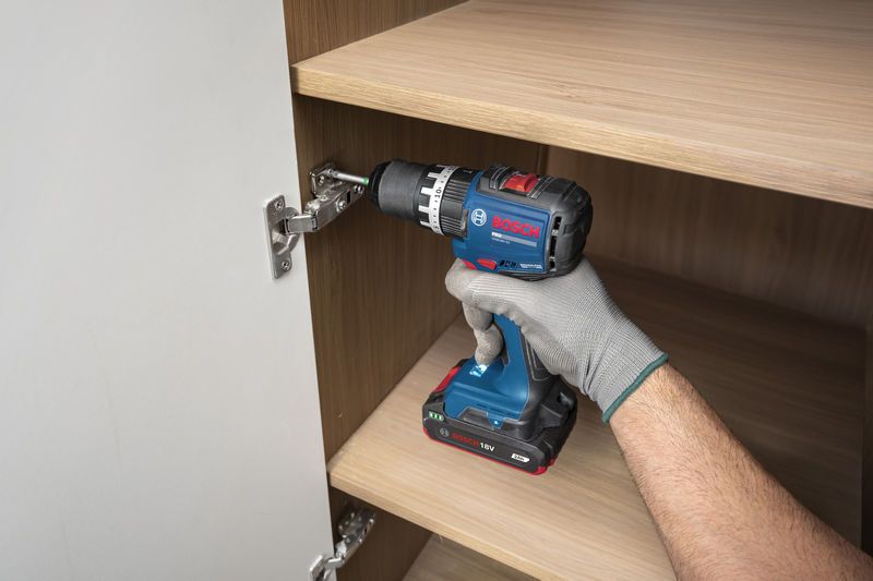 Một người đàn ông mặc đồ bảo hộ đang sử dụng máy khoan không dây để lắp đặt bản lề tủ.