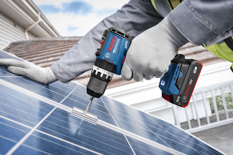 Một người đàn ông mặc đồ bảo hộ đang dùng máy khoan không dây để cố định giá đỡ vào tấm pin mặt trời.