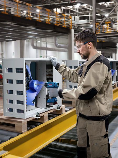 Một người đàn ông mặc đồ bảo hộ đang dùng máy khoan điện để lắp ráp một chiếc quạt công nghiệp.