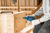 Một người mặc đồ bảo hộ đang khoan vào một thanh gỗ bằng máy khoan điện.