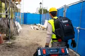 Một người mặc trang phục bảo hộ lao động đang mang ba lô và hộp đựng dụng cụ của nhà thầu tại công trường xây dựng.