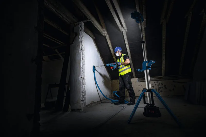 Một người mặc đồ bảo hộ đang khoan vào tường, được chiếu sáng bằng đèn công trường không dây.