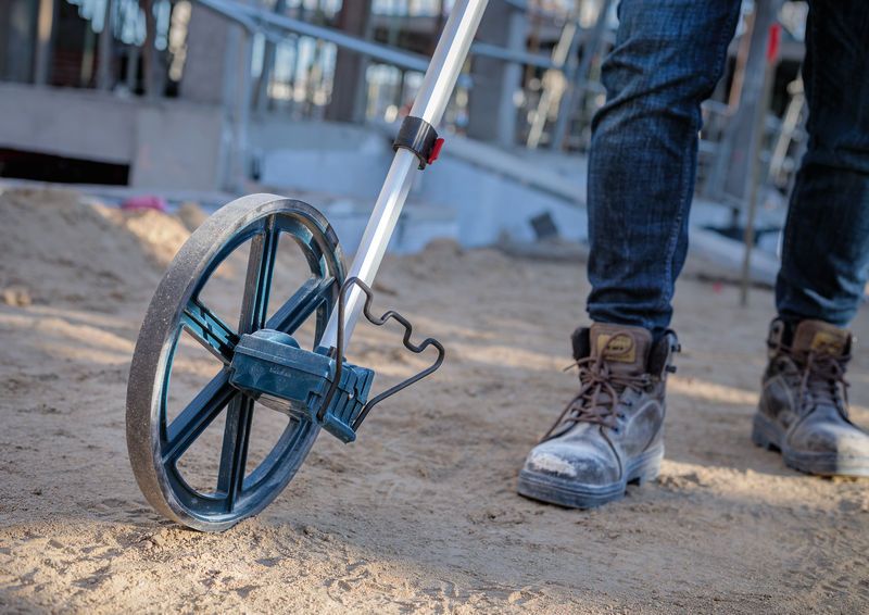 Một người đi ủng lao động đang sử dụng bánh xe đo trên công trường xây dựng đầy cát.