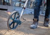 Một người đi ủng lao động đang sử dụng bánh xe đo trên công trường xây dựng đầy cát.