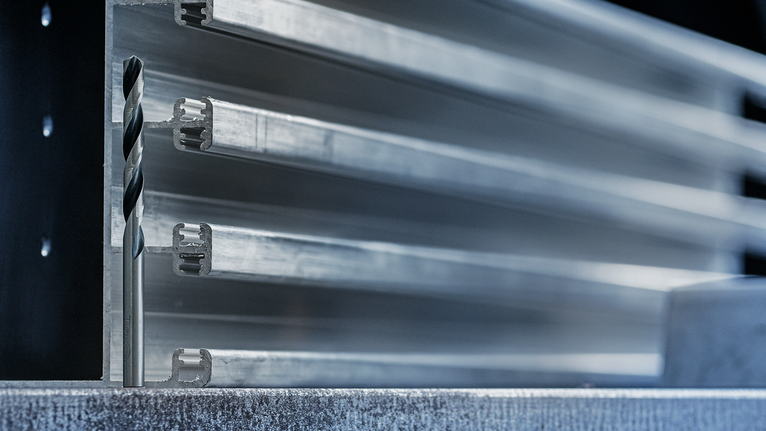 Mũi khoan được đặt thẳng đứng trên bề mặt kim loại ở phía trước các thanh kim loại xếp chồng lên nhau.