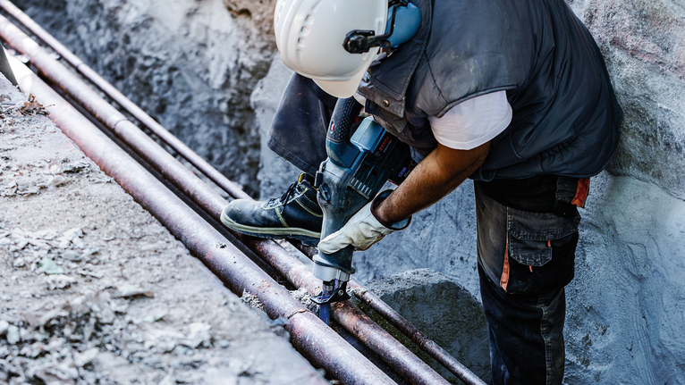 Một người đàn ông mặc đồ bảo hộ đang sử dụng dụng cụ điện để cắt ống kim loại ngoài trời.