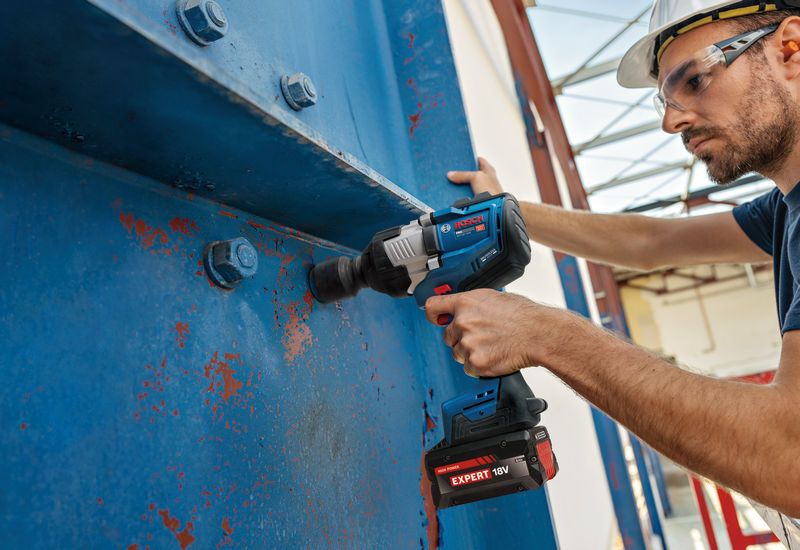 Một người đàn ông mặc đồ bảo hộ đang dùng máy siết bu lông để siết chặt các bu lông trên một thanh dầm kim loại lớn.
