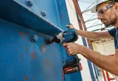 Một người đàn ông mặc đồ bảo hộ đang dùng máy siết bu lông để siết chặt các bu lông trên một thanh dầm kim loại lớn.