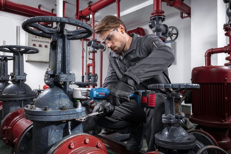 Một người đàn ông mặc đồ bảo hộ đang sử dụng máy siết bu lông không dây để siết chặt van trong môi trường công nghiệp.