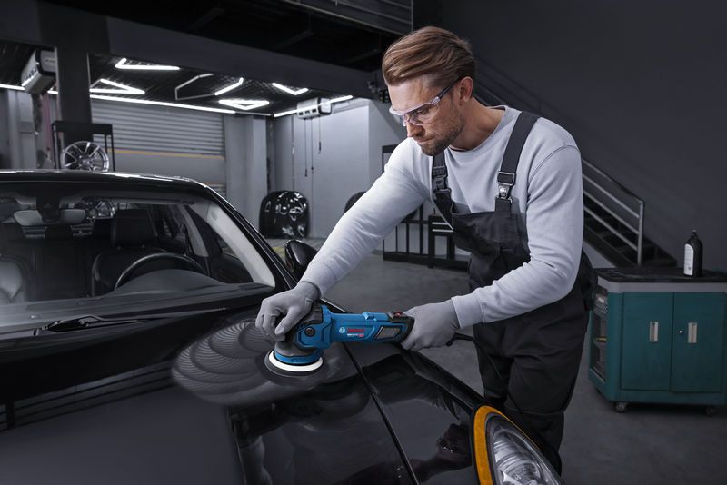 Một người đàn ông mặc đồ bảo hộ đang dùng máy đánh bóng điện để đánh bóng nắp ca-pô xe màu đen.