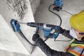 Một người đàn ông mặc đồ bảo hộ đang dùng máy khoan búa xoay và máy hút bụi để khoan vào bê tông.