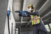 Một công nhân mặc đồ bảo hộ đang dùng máy khoan búa xoay để khoan vào bức tường bê tông.