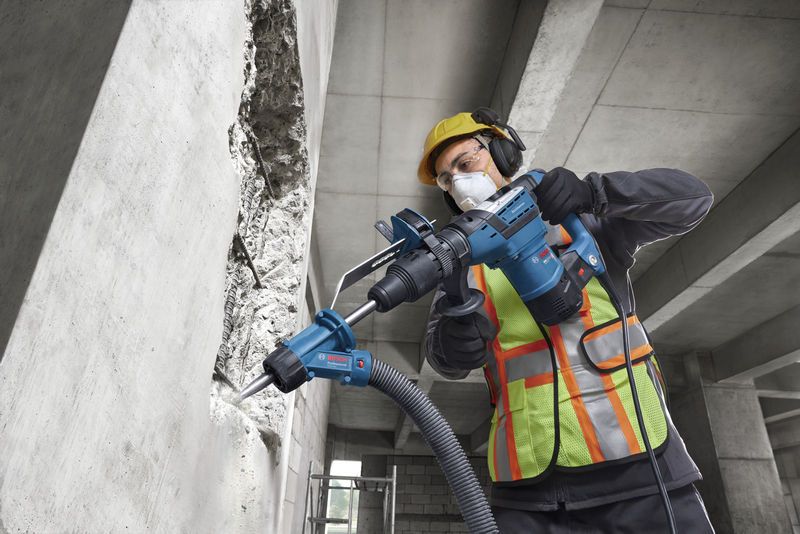 Một công nhân mặc đồ bảo hộ đang sử dụng máy khoan búa xoay để khoan vào tường bê tông.