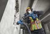 Một công nhân mặc đồ bảo hộ đang sử dụng máy khoan búa xoay để khoan vào tường bê tông.