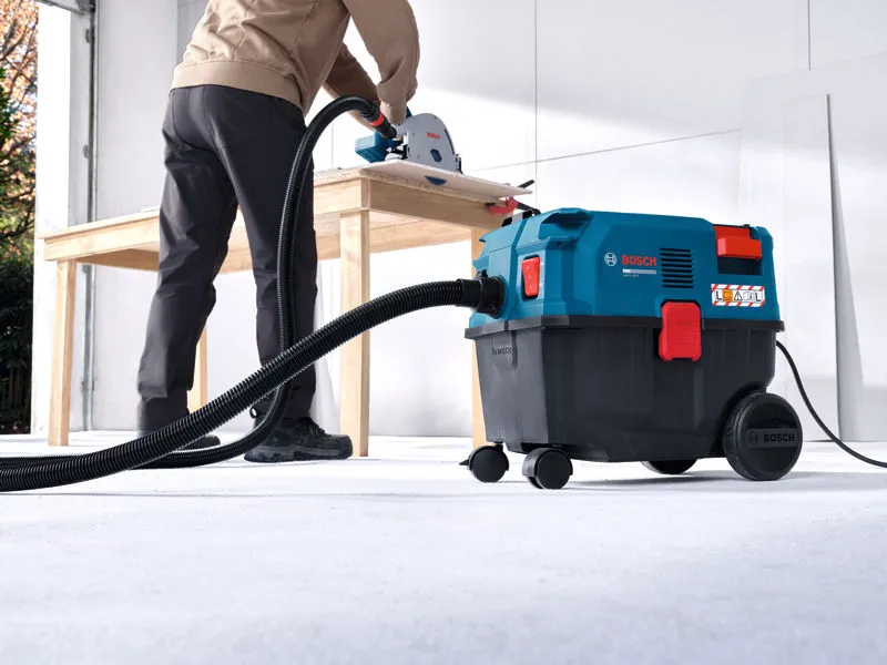 Một người đàn ông mặc đồ bảo hộ đang sử dụng máy hút bụi trong khi cắt gỗ trên bàn.