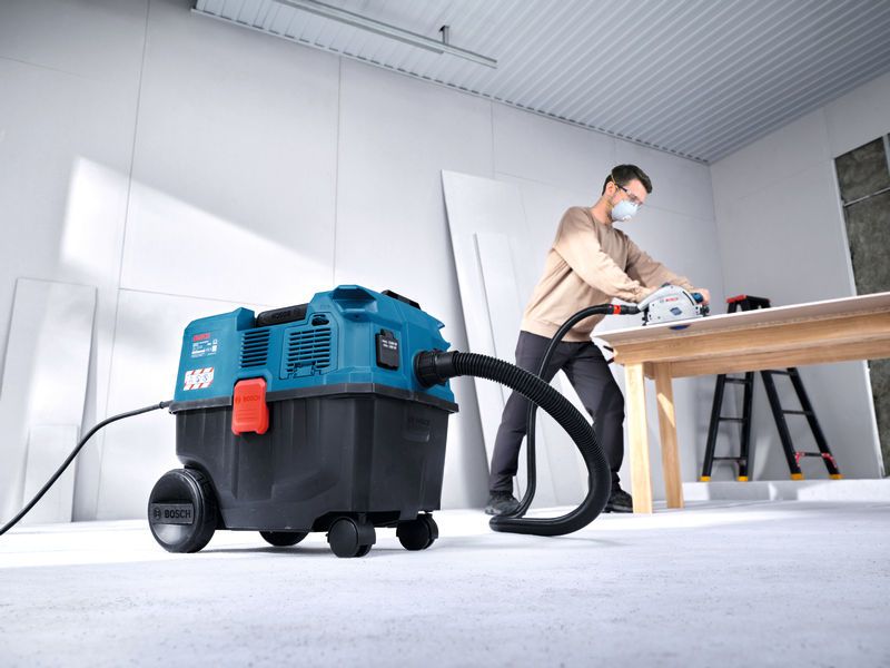 Một người đàn ông mặc đồ bảo hộ đang sử dụng máy hút bụi đa năng để cắt gỗ trên bàn.