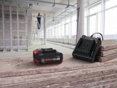 Pin sạc và bộ sạc được đặt trên một bề mặt gỗ, phía sau là một người đang làm việc trên thang.