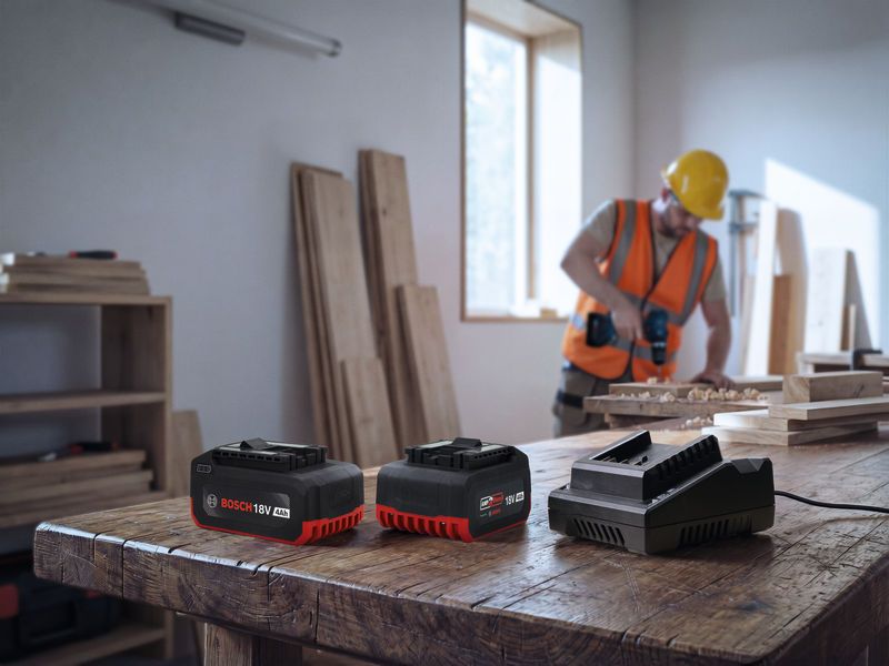 Một người đàn ông mặc đồ bảo hộ đang khoan gỗ trong một xưởng, xung quanh là các bộ pin dụng cụ điện đặt trên bàn.