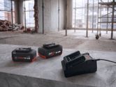 Hai cục pin sạc dùng cho dụng cụ và một bộ sạc được đặt trên bề mặt bê tông tại một công trường xây dựng.