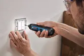 Một người đàn ông mặc đồ bảo hộ đang dùng tua vít không dây để lắp đặt công tắc điện trên tường.