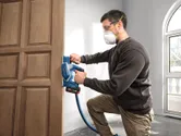 Một người đàn ông mặc đồ bảo hộ đang sử dụng máy bào không dây trên một cánh cửa gỗ.