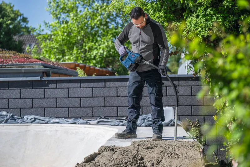 Một người đàn ông mặc đồ bảo hộ đang sử dụng dụng cụ điện để đầm nén bê tông ngoài trời.