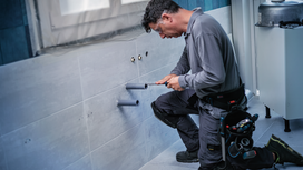 Một người đàn ông mặc đồ bảo hộ đang lắp đặt đường ống trong bức tường lát gạch của phòng tắm.