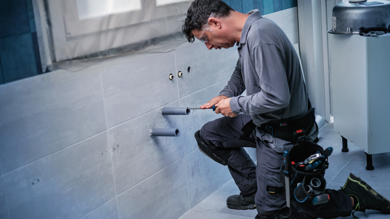 Một người đàn ông mặc đồ bảo hộ đang lắp đặt đường ống trong bức tường lát gạch của phòng tắm.