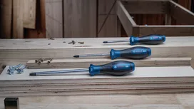 Ba chiếc tua vít cán xanh nằm trên những tấm ván gỗ xếp chồng lên nhau, bên cạnh một vài con ốc vít.