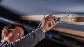 Một người đàn ông mặc đồ bảo hộ đang dùng dụng cụ cầm tay để bắt vít vào khung trần nhà.