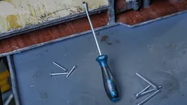 Một chiếc tua vít dài được đặt trên bề mặt kim loại với nhiều ốc vít nằm rải rác.