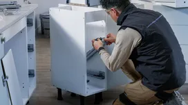 Một người đàn ông mặc đồ bảo hộ đang lắp ray trượt ngăn kéo bên trong một chiếc tủ màu trắng.