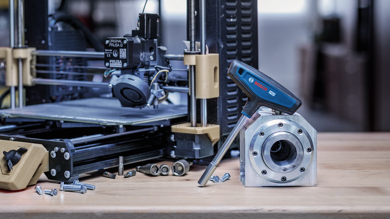 Cờ lê lực điện tử được đặt trên một chi tiết kim loại bên cạnh máy in 3D trên bàn làm việc.