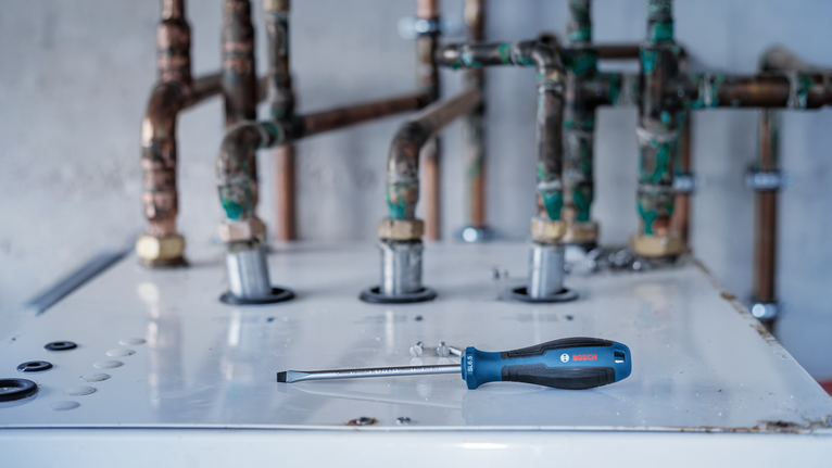 Một chiếc tua vít đầu dẹt nằm trên đỉnh một cái nồi hơi gần một vài đường ống bị rỉ sét.