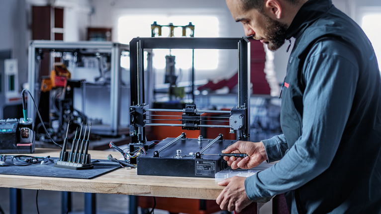 Một người đàn ông mặc đồ bảo hộ đang điều chỉnh máy in 3D trên bàn làm việc.