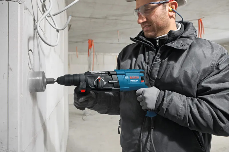 GBH 2-28 DFV Một người mặc đồ bảo hộ đang khoan một lỗ lớn trên bức tường bê tông bằng một dụng cụ điện.