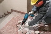 Một người mặc đồ bảo hộ đang sử dụng công cụ phá dỡ để phá vỡ gạch lát sàn.